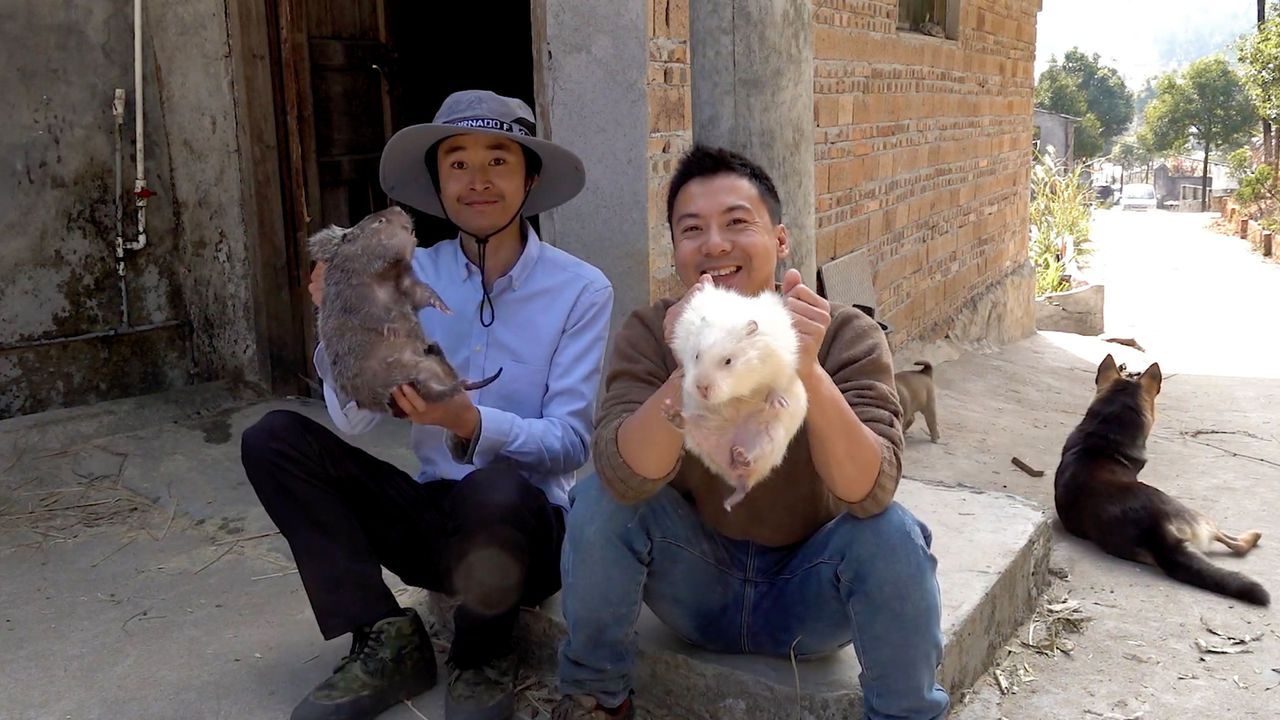 Huanong Brothers sitting in front of a building, holding up Bamboo rats.